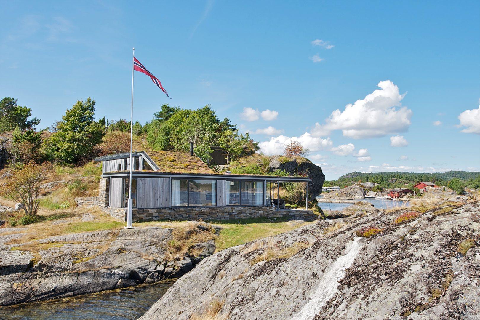 Moderne hytte med flatt tak bak et svaberg, norsk flagg i vinden